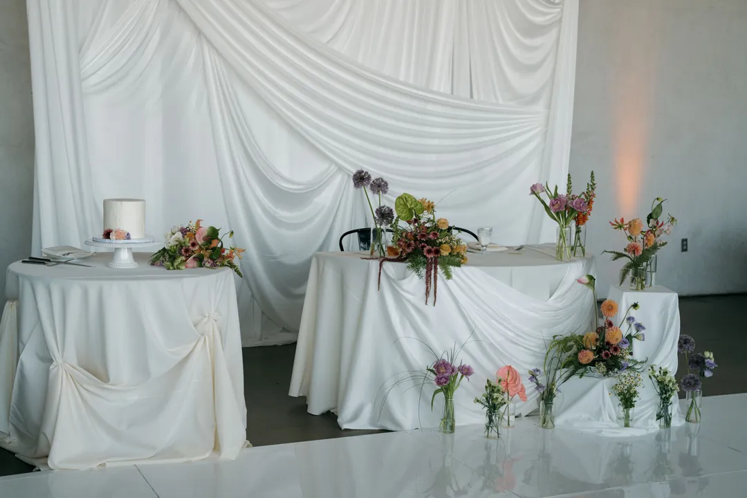 White-draped sweetheart table with colorful flowers and a matching fabric backdrop.