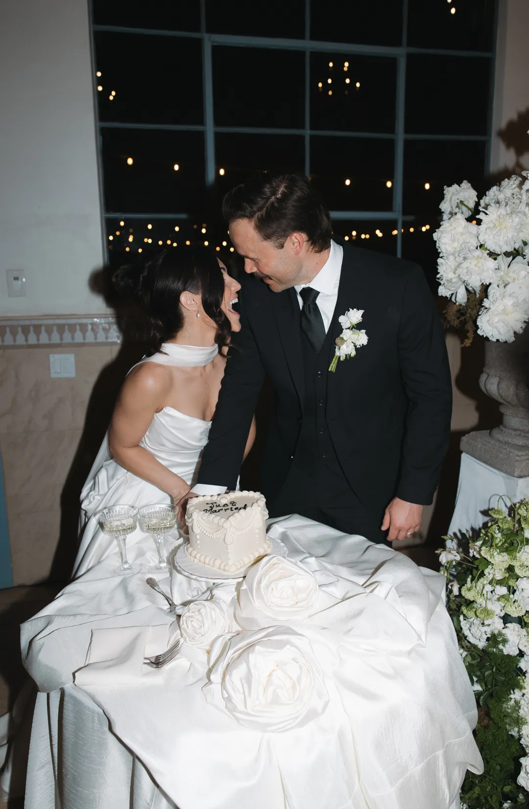 Wedding couple kissing beside their cake at a white-draped table.