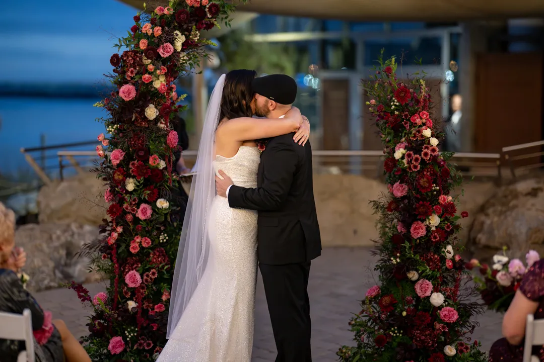 Wedding couple kissing beneath a floral arch beside the water.