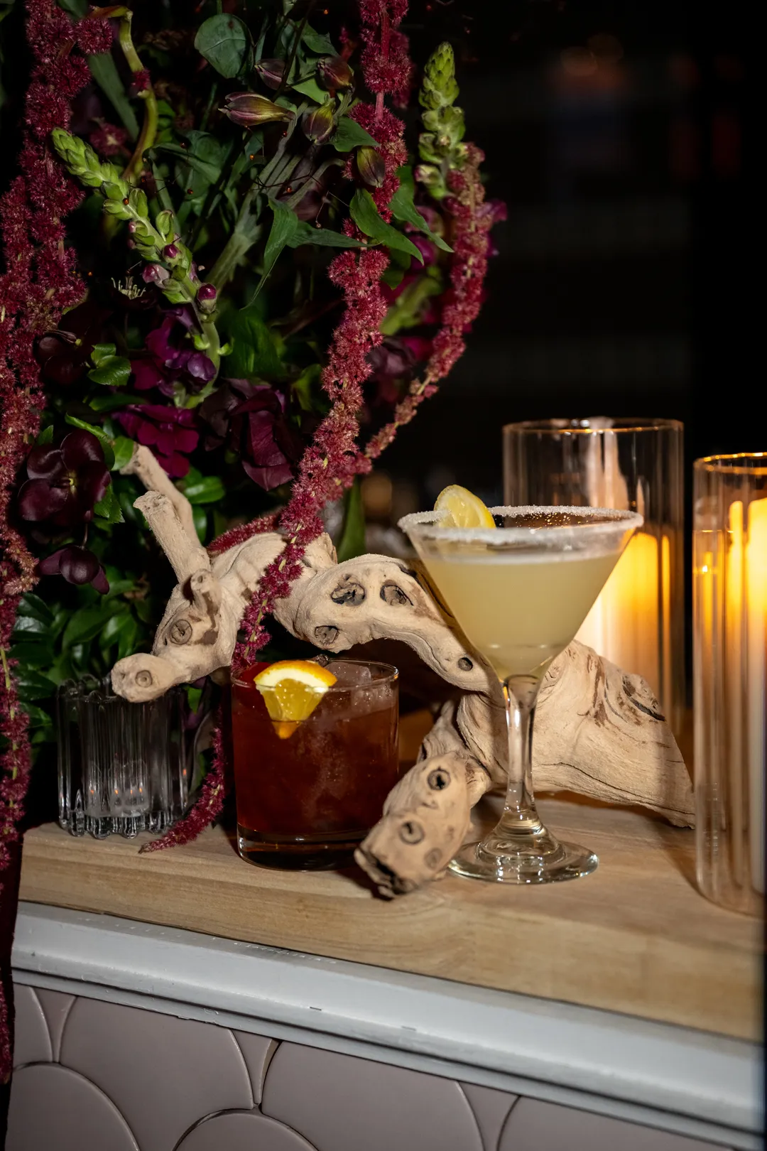 Cocktails displayed beside decorative wood, flowers and candles.