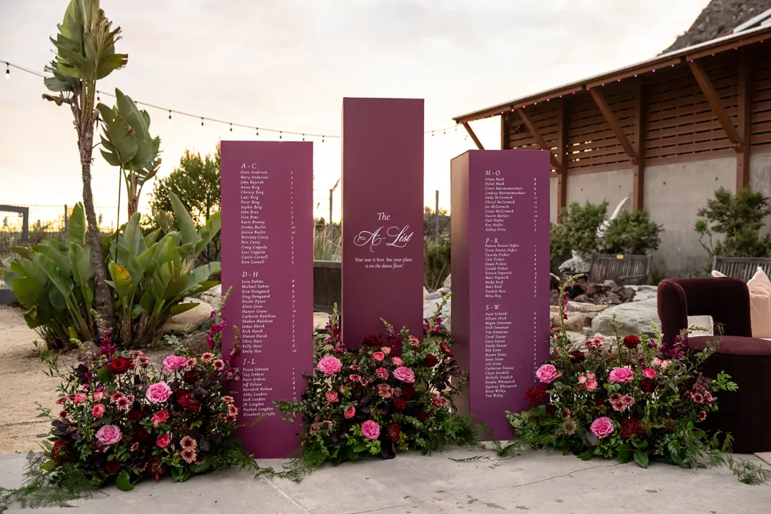 Plum-colored wedding seating chart surrounded by pink and burgundy flowers.