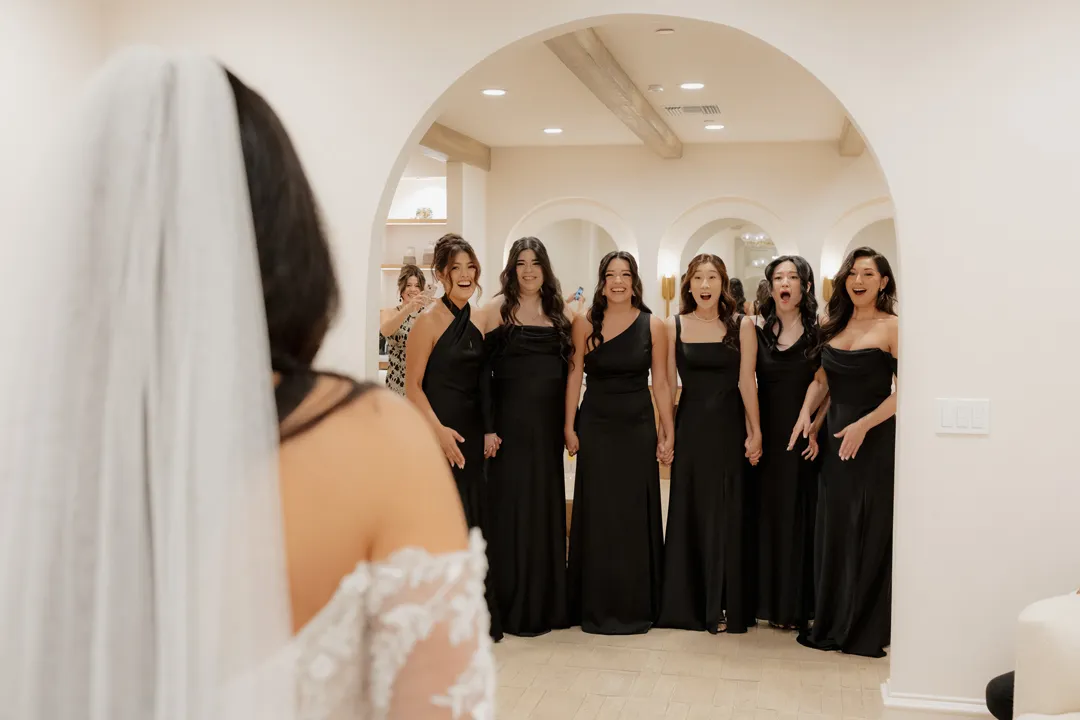 Bridesmaids in black dresses reacting to the bride's wedding gown.