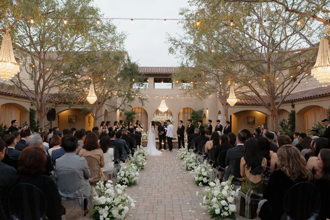 Wedding ceremony beneath trees and chandeliers, with white flowers lining the aisle.