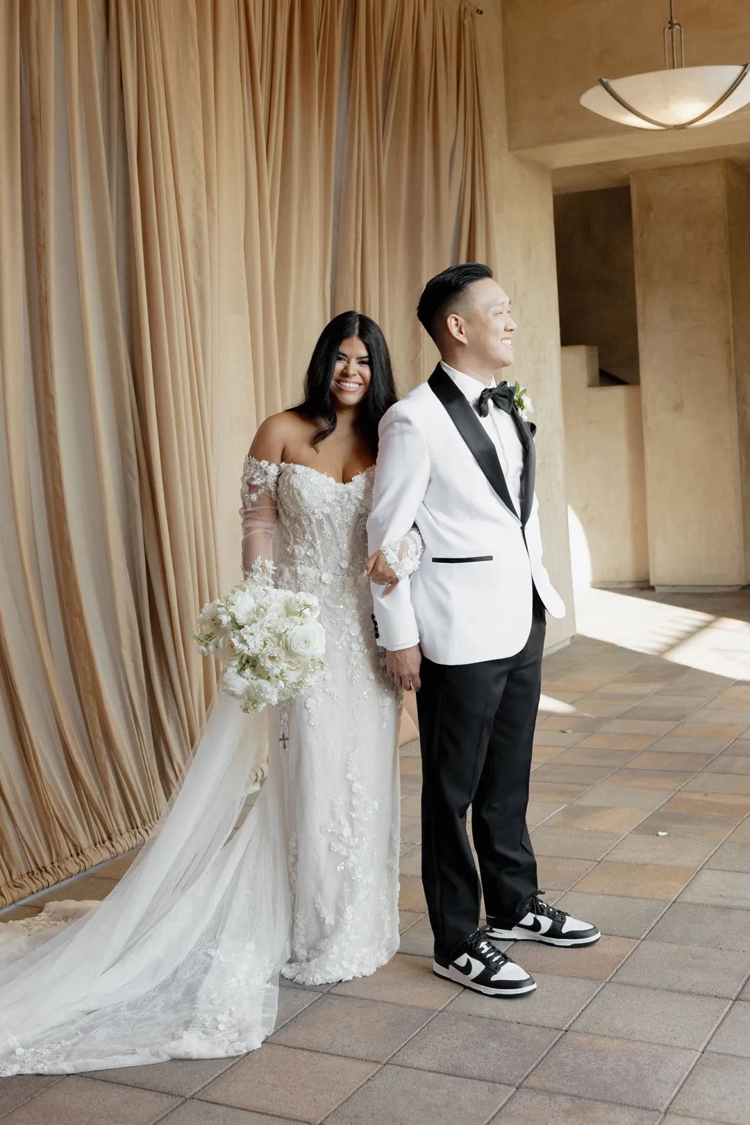 Wedding couple standing together with a white bouquet beside a draped doorway.