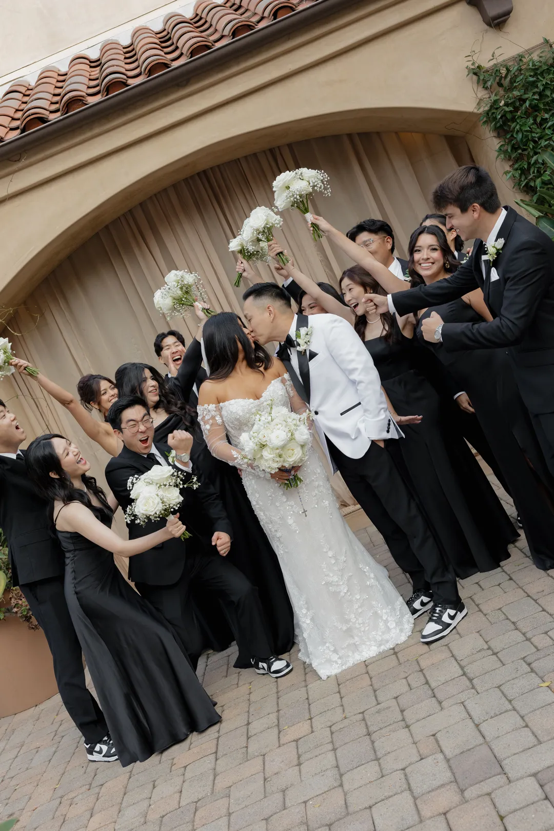 Wedding party cheering around the couple beneath a courtyard arch.