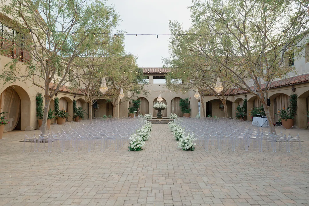 Courtyard ceremony setup with clear chairs and a white floral aisle.