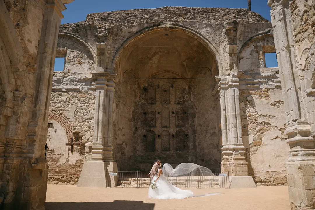 Wedding couple framed by a tall stone arch, with the bride's veil flowing behind her.