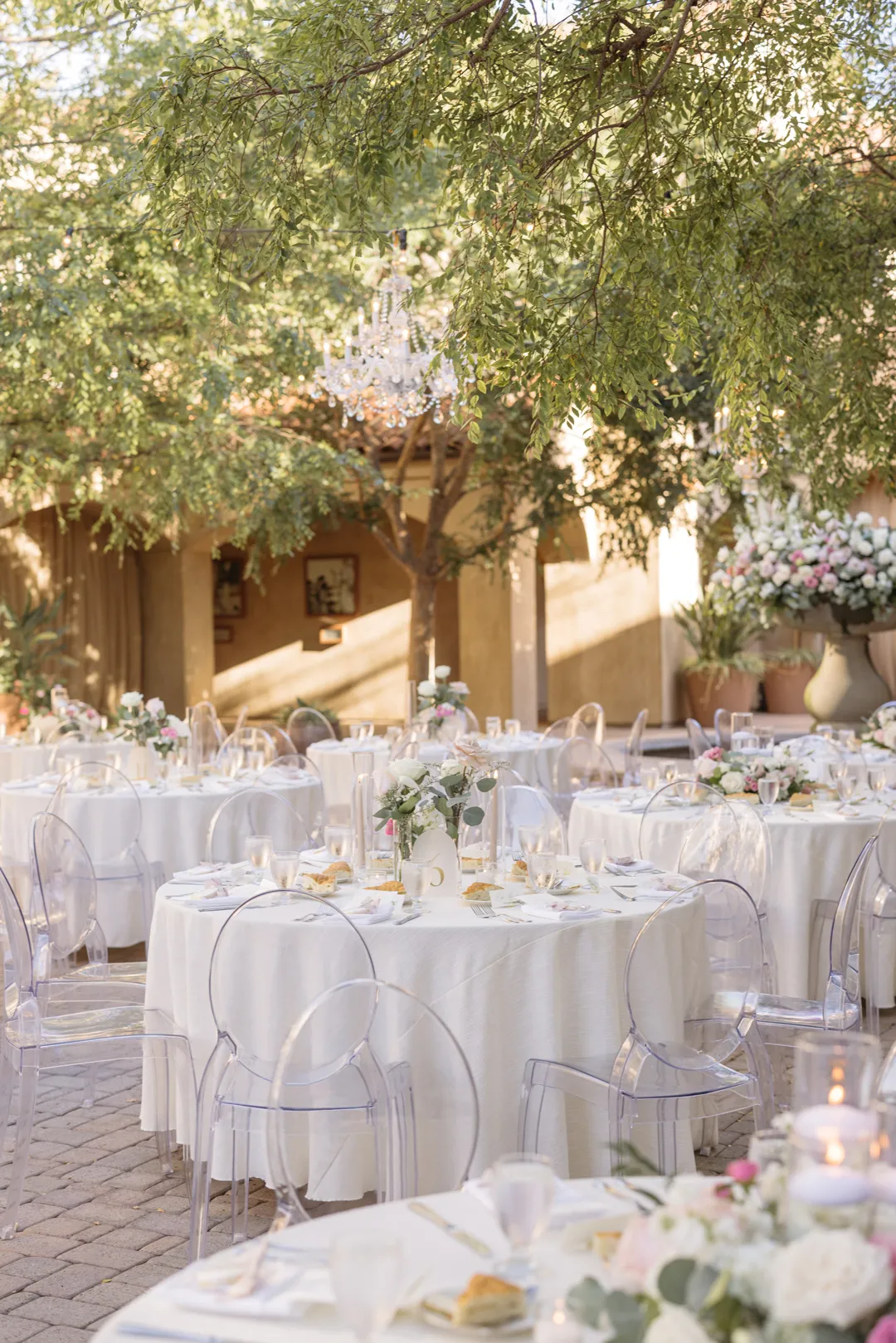 Reception tables with clear chairs and blush floral centerpieces beneath courtyard trees.