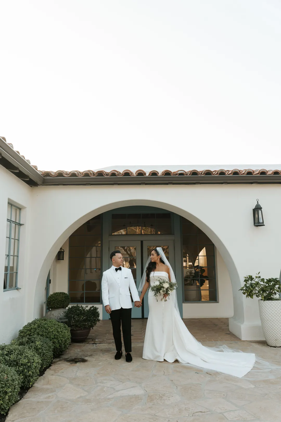 Wedding couple holding hands beneath a white stucco arch.