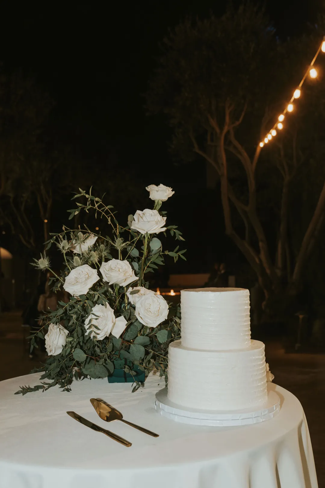 Two-tier white wedding cake beside white roses and greenery.