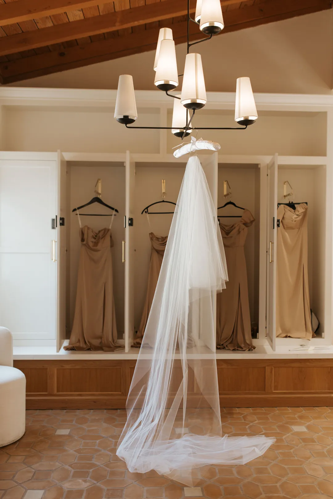 Wedding veil hanging beneath pendant lights in a dressing room with champagne-colored dresses.