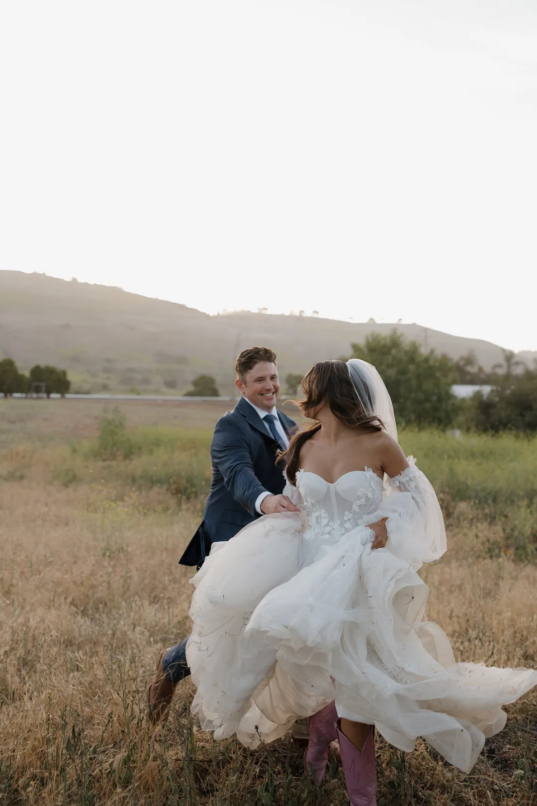 Wedding couple running hand in hand through a grassy field.