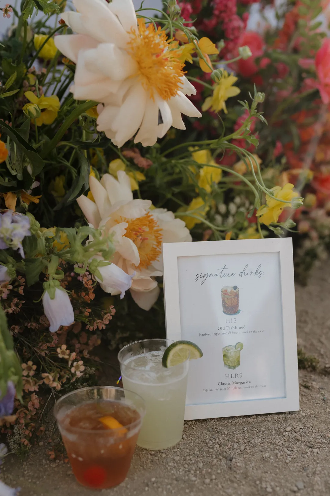 Two signature cocktails beside a framed drink menu and colorful flowers.