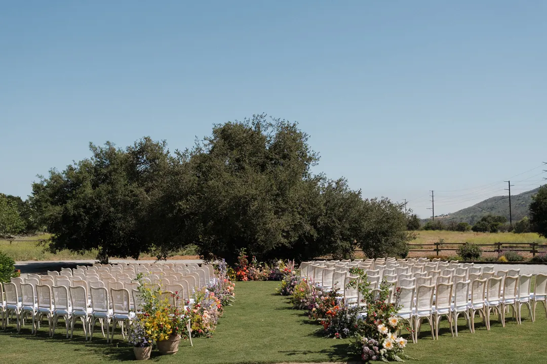 Outdoor ceremony lawn with white chairs and a colorful flower-lined aisle.