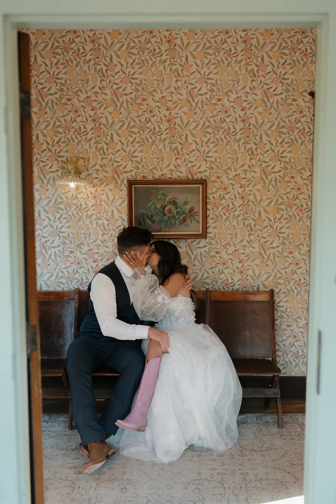 Wedding couple kissing on a bench in a room with floral wallpaper.