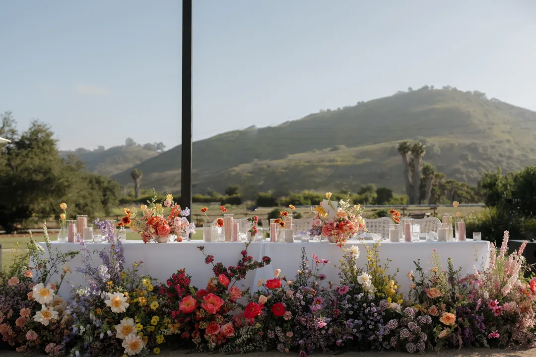 Sweetheart table surrounded by colorful flowers with a green hillside behind it.