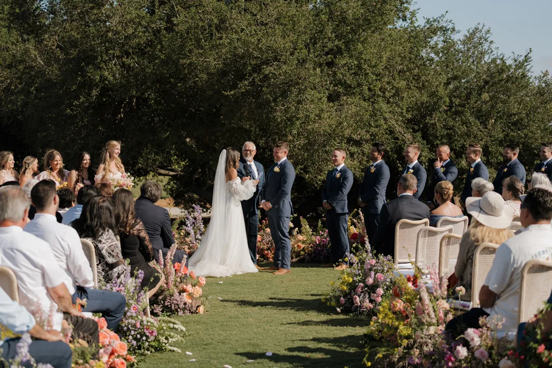 Wedding couple facing each other at the end of the flower-lined aisle.