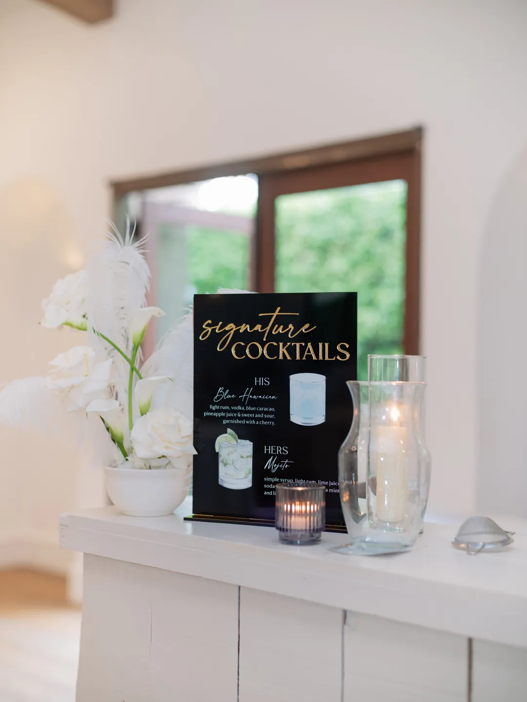 Black and gold signature cocktail menu beside white flowers and candles.