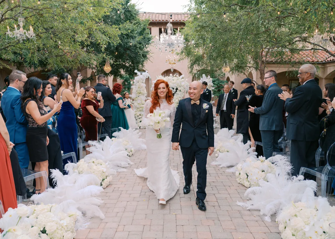 Newlyweds walking down an aisle lined with white flowers and feathers.