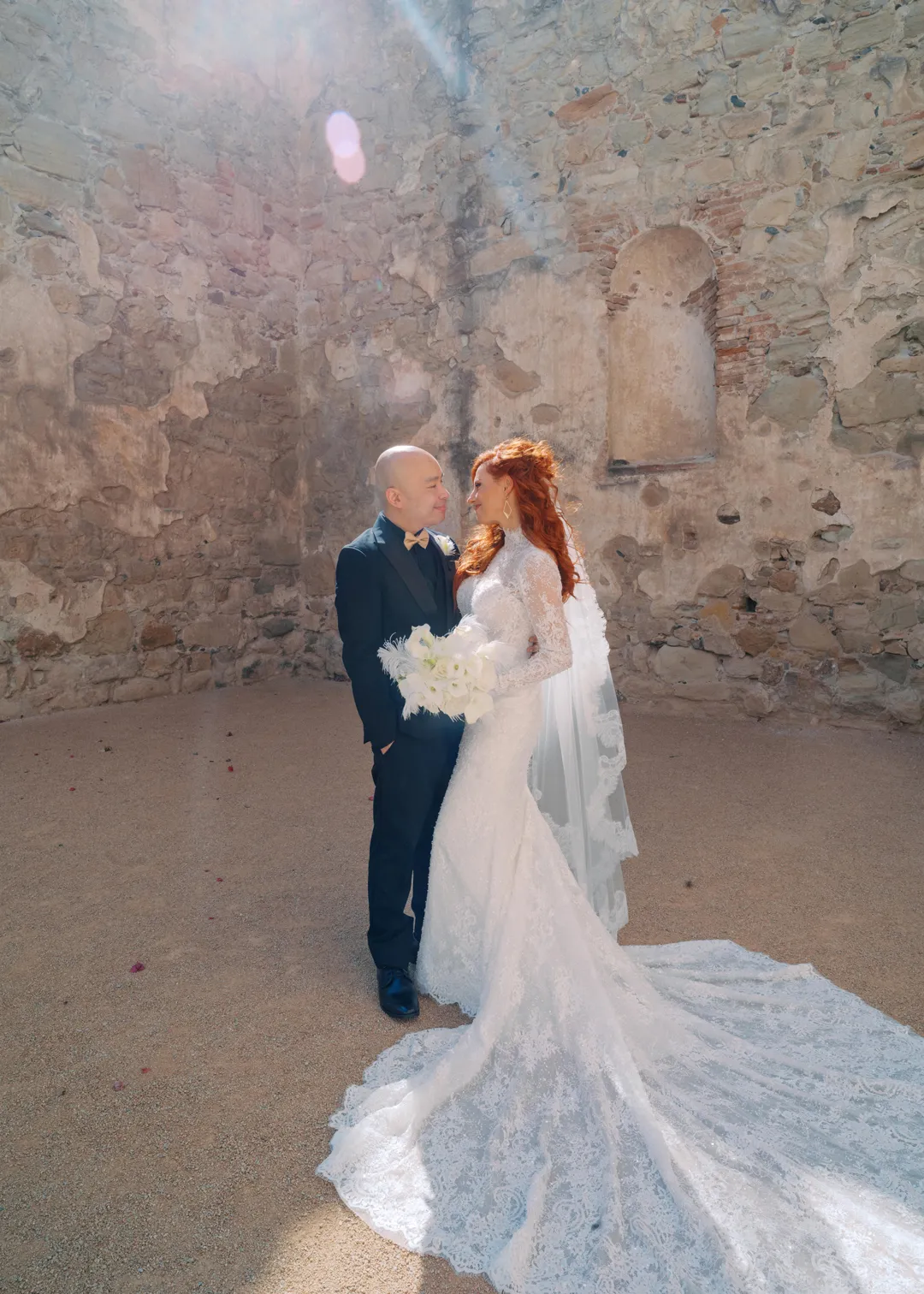 Wedding couple posing beside an old stone wall with the bride's lace train spread behind her.