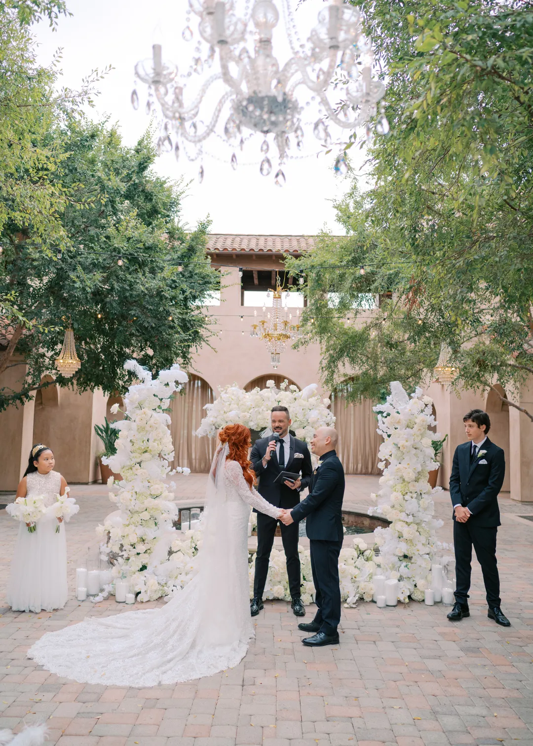 Wedding couple exchanging vows before a white-flower-covered courtyard fountain.