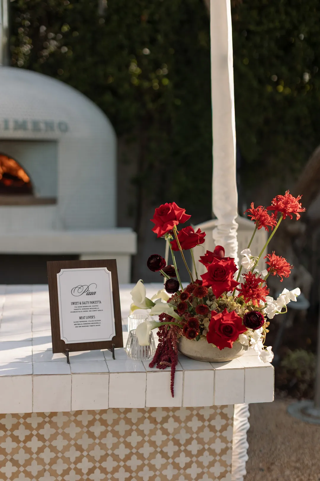 Red flowers and a small menu displayed beside the outdoor pizza oven.