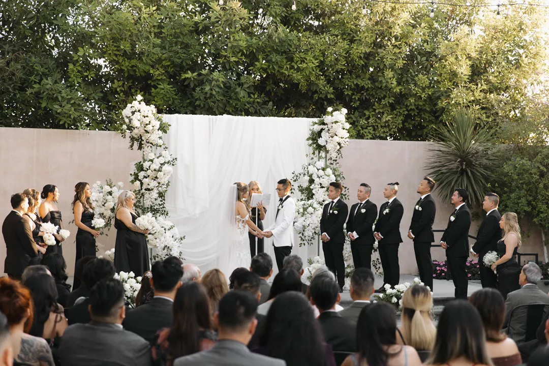 Wedding couple exchanging vows before a white floral ceremony backdrop.