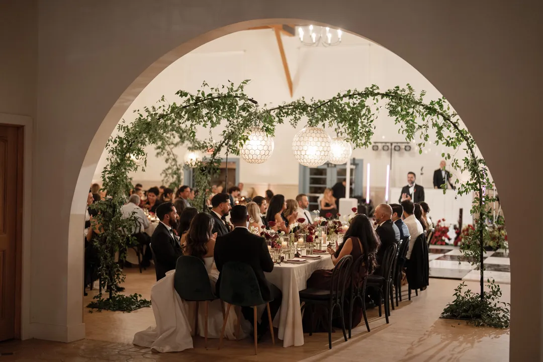 Wedding guests seated beneath a greenery arch, viewed through an arched doorway.