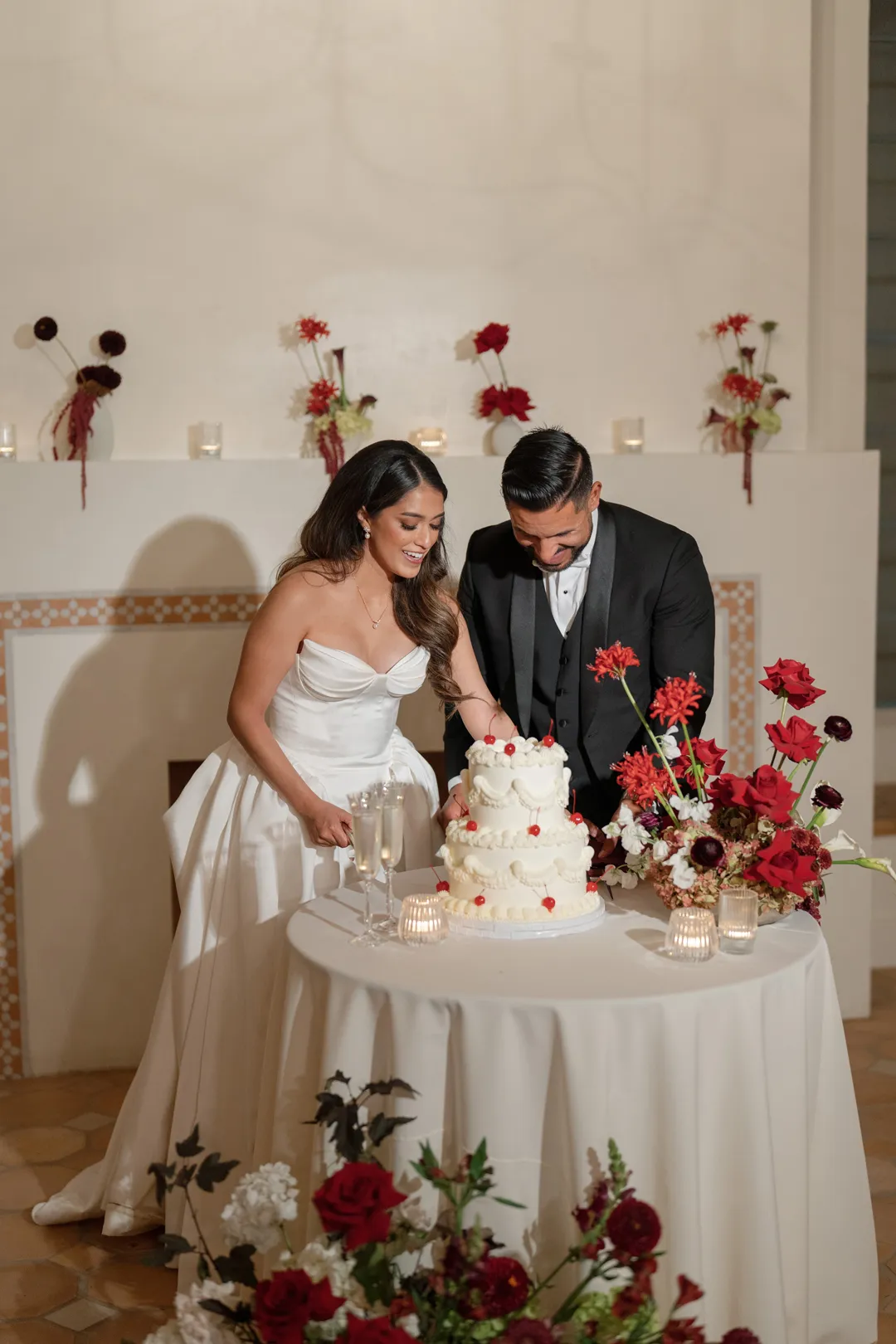 Wedding couple cutting a white tiered cake beside red flowers.