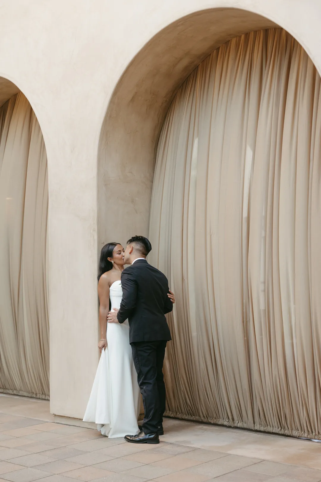 Wedding couple kissing beneath a tall curtained arch.