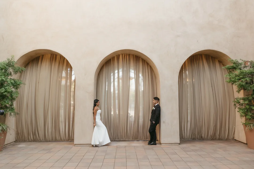 Wedding couple facing each other in front of three courtyard arches.