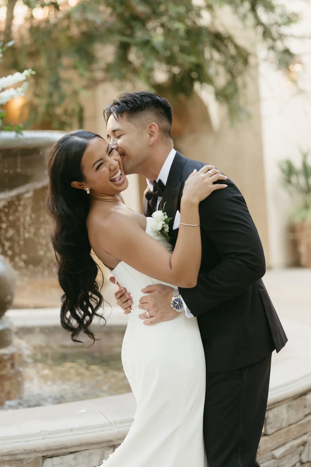 Groom kissing the bride's cheek beside the courtyard fountain.