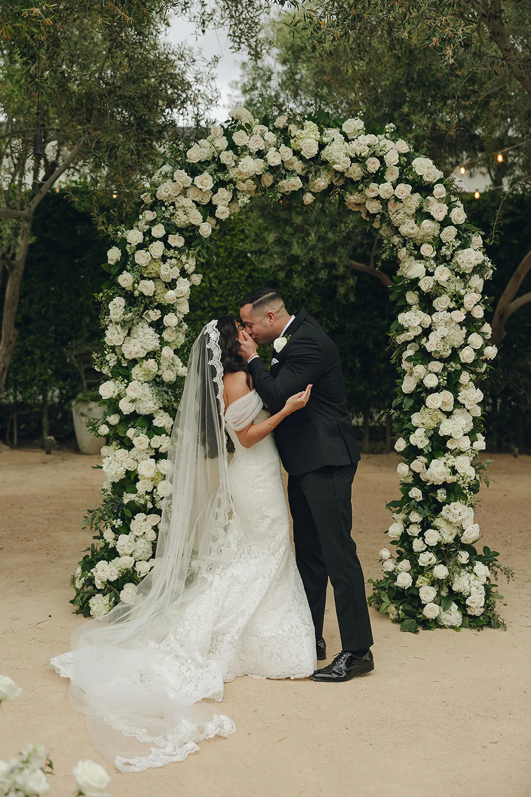 Wedding couple kissing beneath their white floral arch.