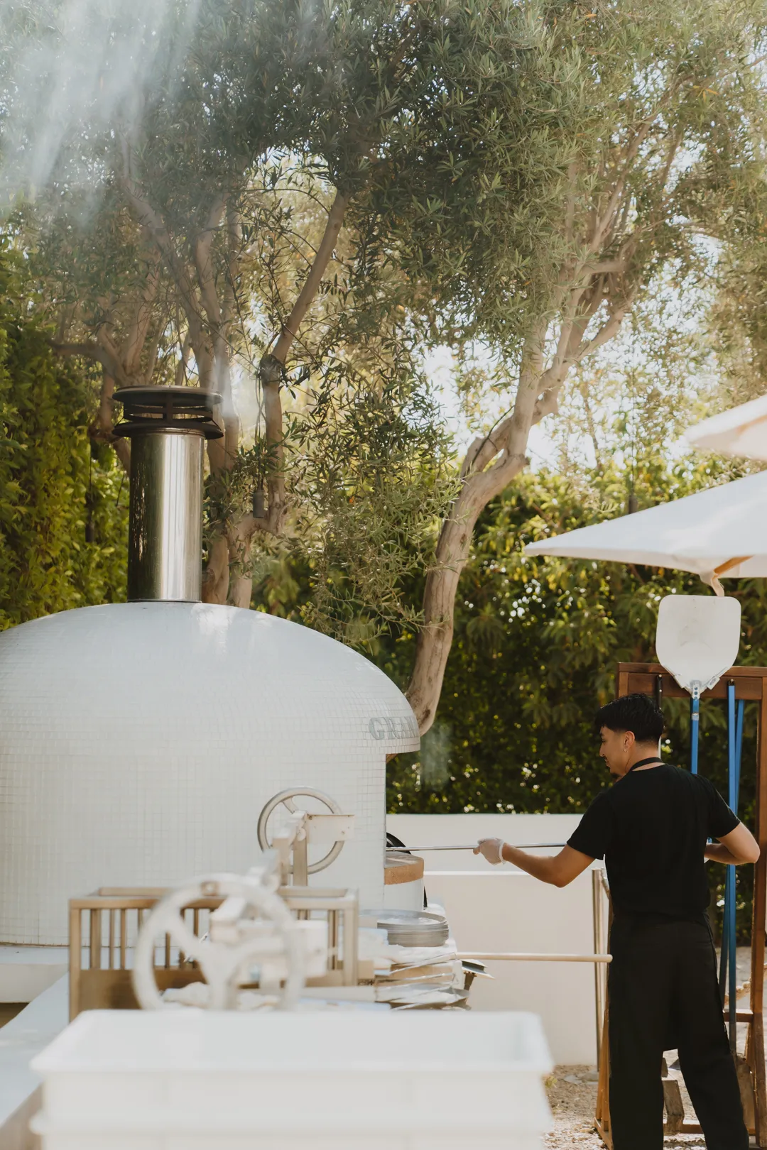 Chef tending the outdoor pizza oven beneath olive trees.