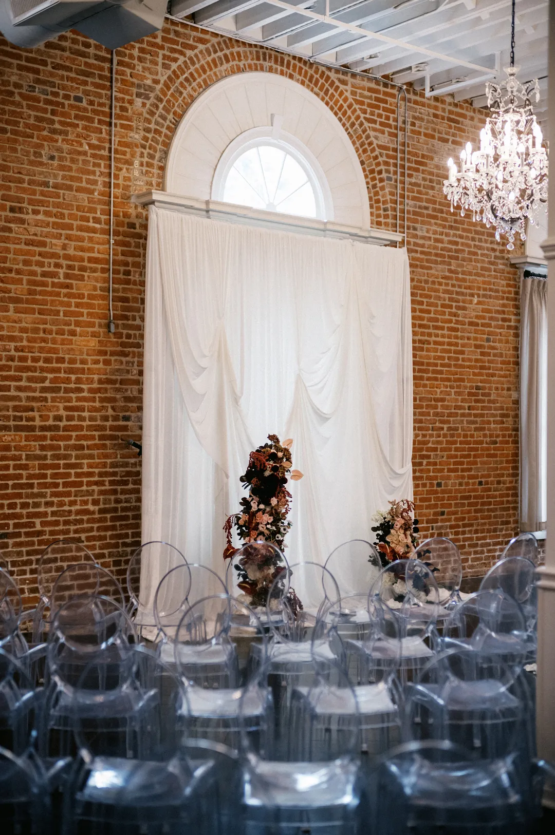 Ceremony seating with clear chairs beneath a tall curtained arch.