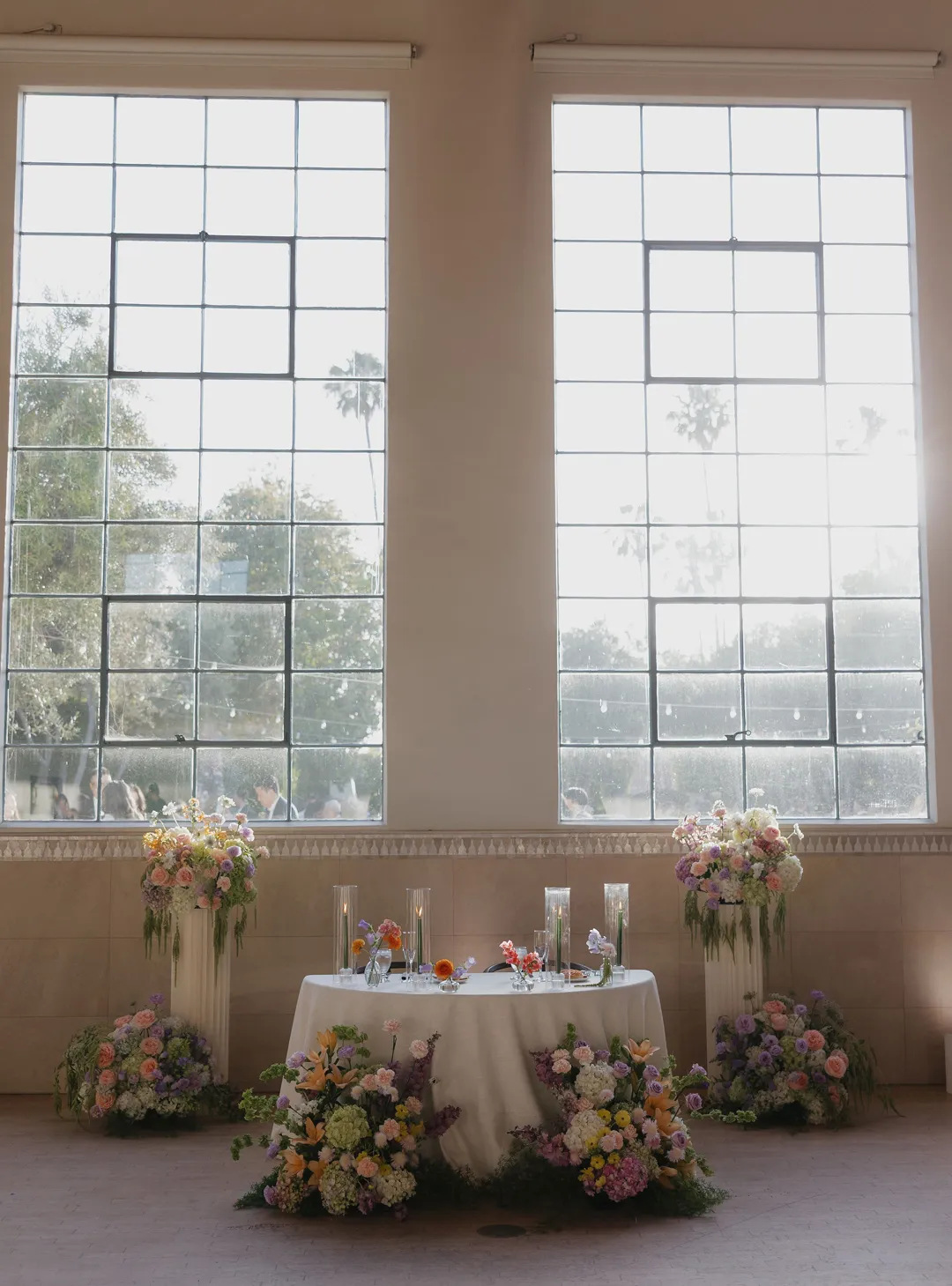 Sweetheart table framed by pastel flowers beneath tall windows.