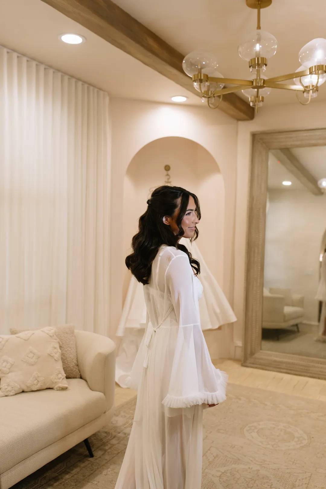 Bride in a sheer white robe beside a lounge sofa.