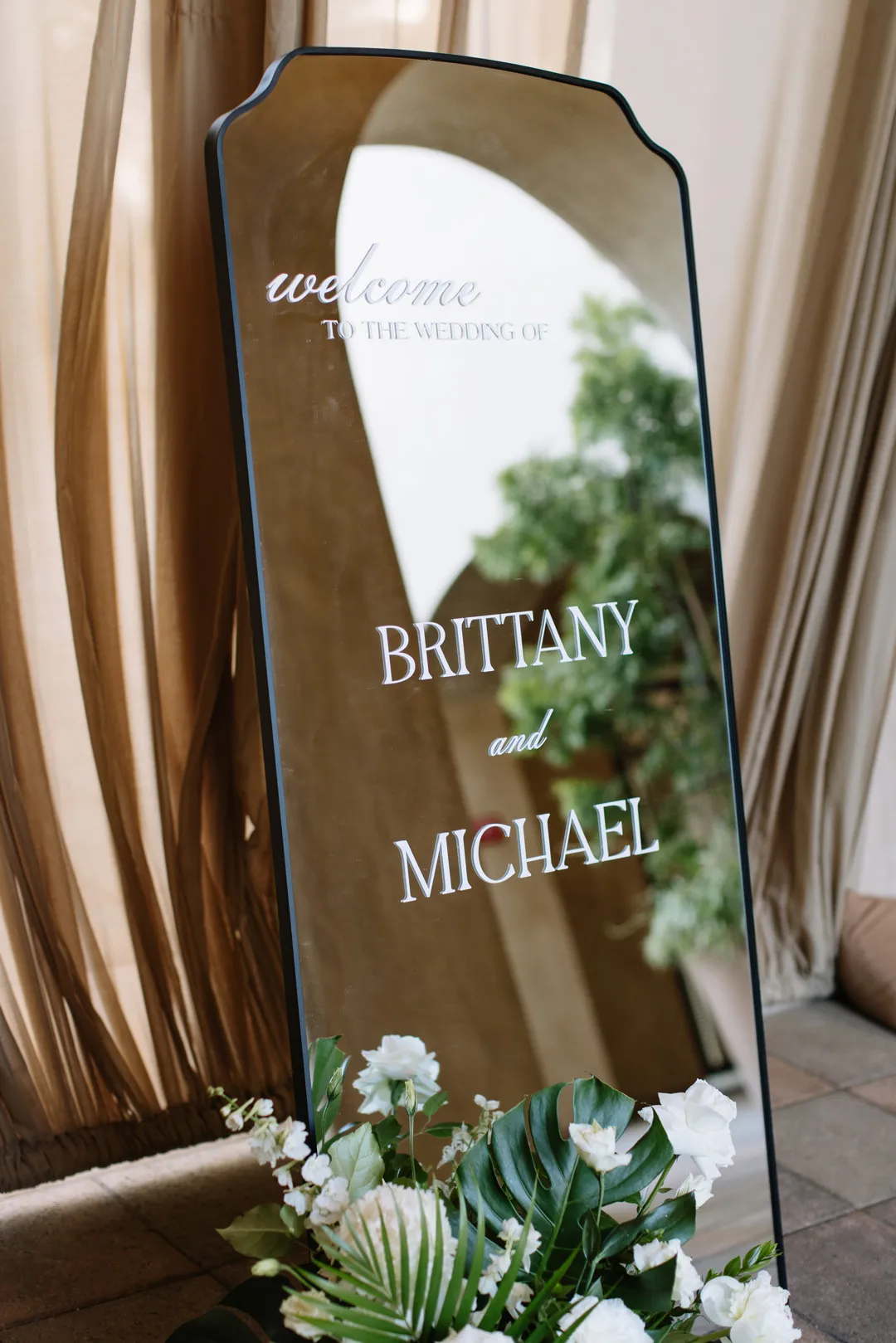 Mirrored wedding welcome sign beside white flowers and tropical greenery.