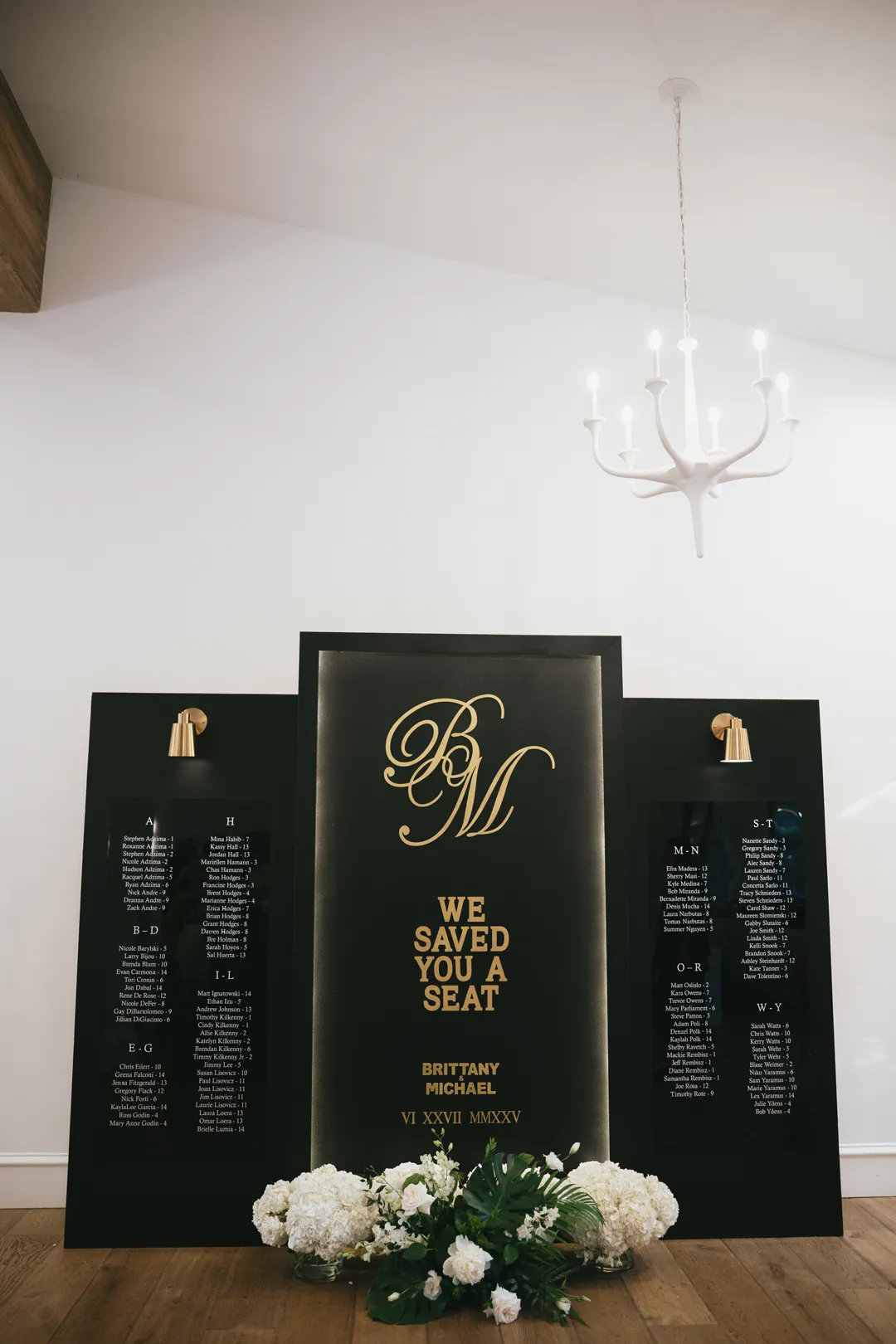 Black-and-gold wedding seating chart with white flowers at its base.