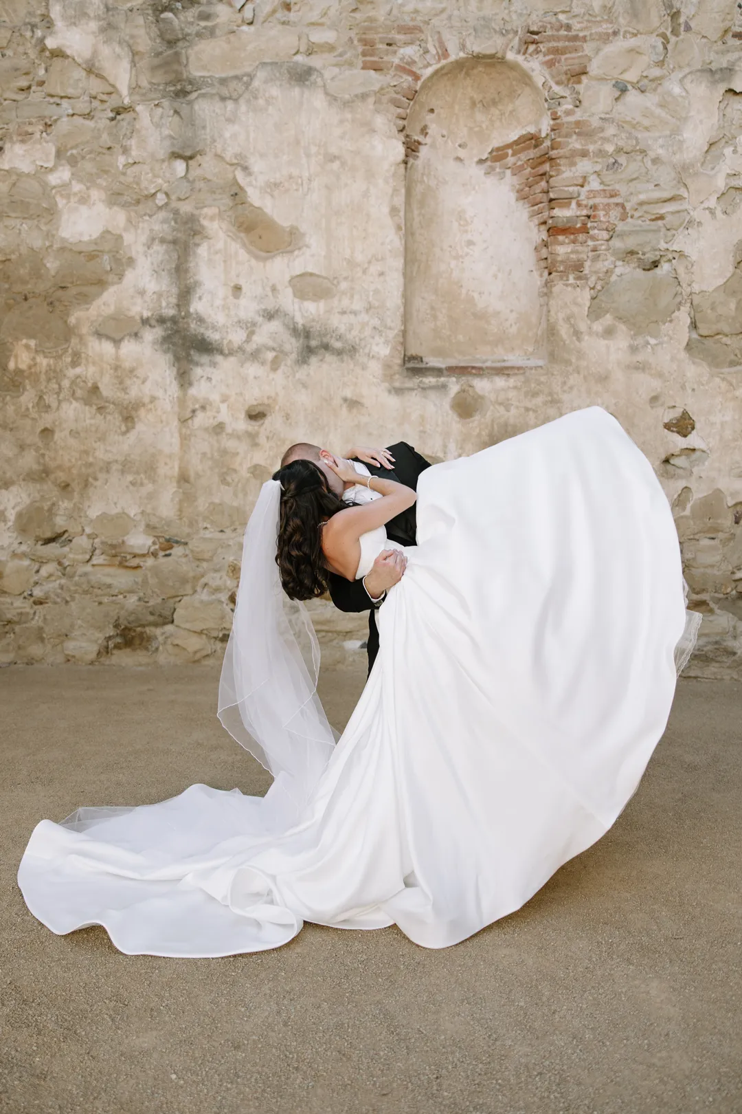 Groom dipping the bride beside a weathered stone wall.