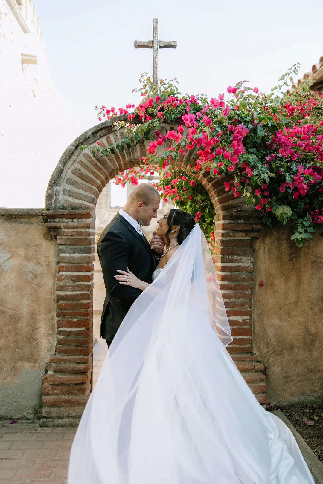 Wedding couple kissing beneath a flower-covered brick arch.