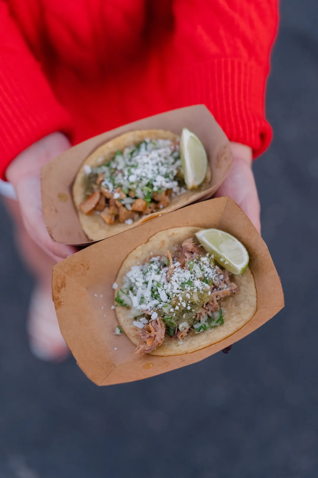 Two tacos in paper trays with lime wedges and crumbled topping.