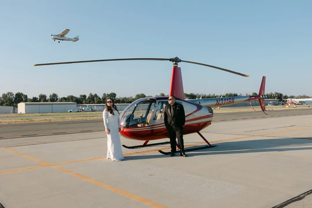 Wedding couple beside a red helicopter as an airplane passes overhead.