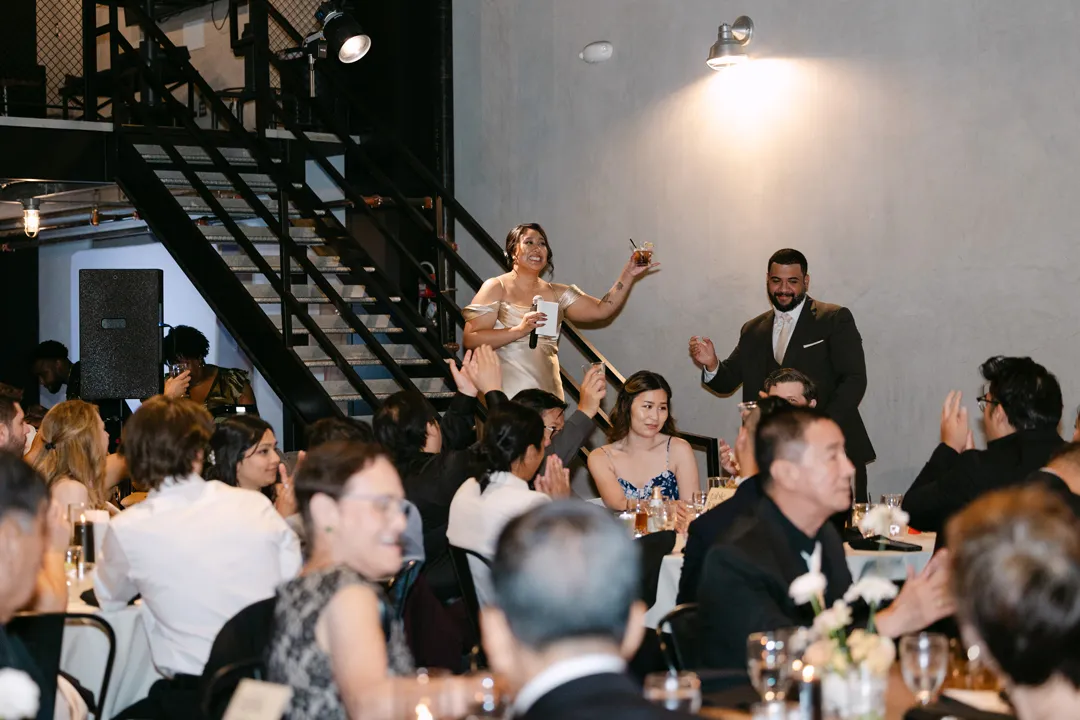 Wedding couple raising glasses among seated reception guests.
