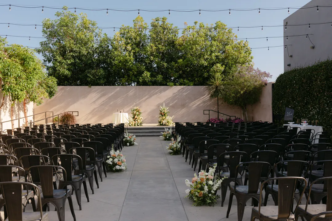 Outdoor ceremony aisle lined with pale flowers and black chairs.