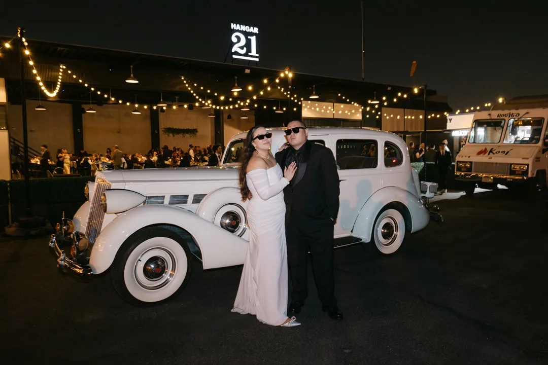 Wedding couple posing beside a vintage white car outside Hangar 21 at night.