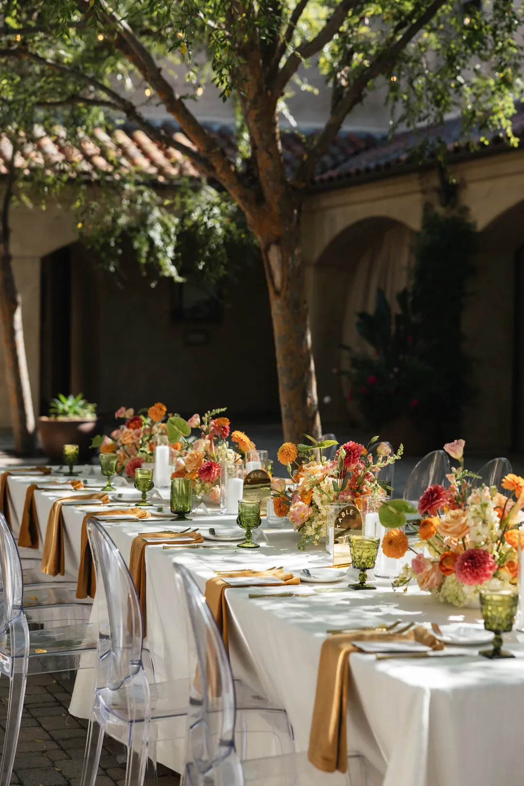 Long reception table with golden napkins, flowers and clear chairs.