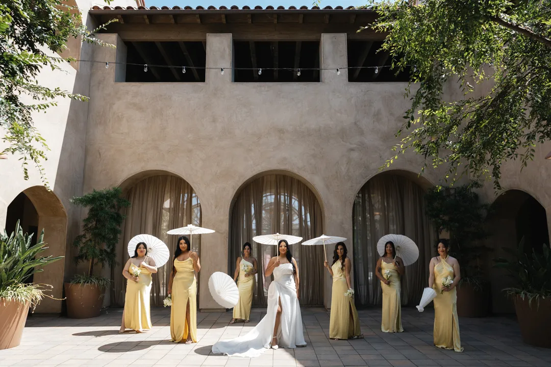 Bride and bridesmaids in yellow dresses holding white parasols beside stucco arches.