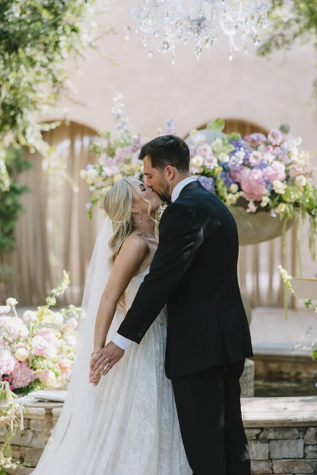 Newlyweds kissing beside pastel flowers and a crystal chandelier.