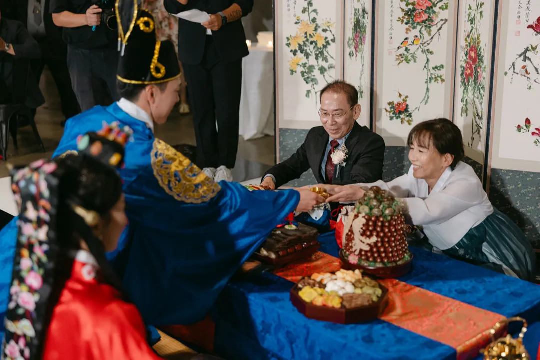 Couple in colorful ceremonial clothing presenting a cup across a decorated table.